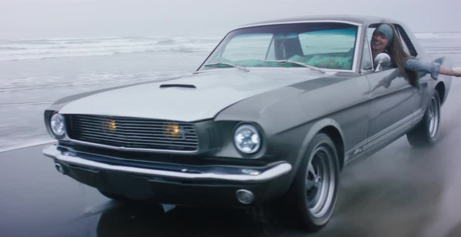 A classic grey Ford Mustang cruises along the wet sand of a foggy coastline as a happy young woman enjoys the ride