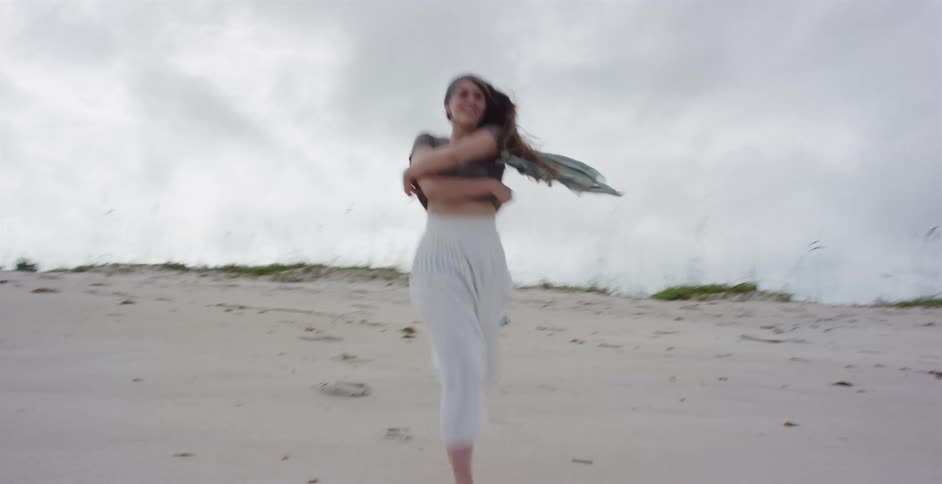 A carefree young woman runs joyfully in slow motion on a vast sand dune, throwing a scarf to the wind under an overcast sky