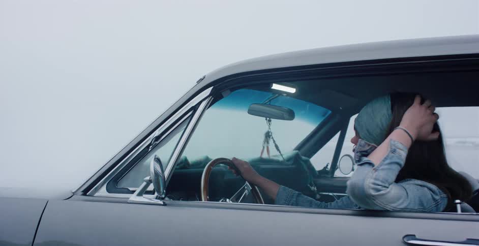 A young woman drives her classic silver car along a foggy coastline and then slowly drives away