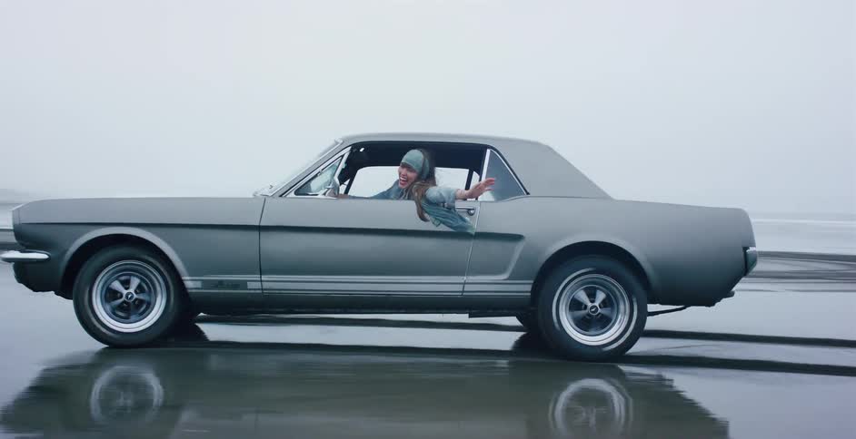 A happy woman hangs out the window of a classic gray Mustang as it drives along a wet beach in slow motion
