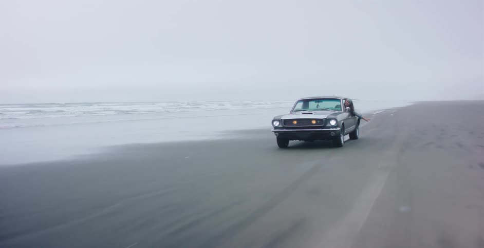 A classic car drives along a misty beach as a person leans out of the window to enjoy the ride