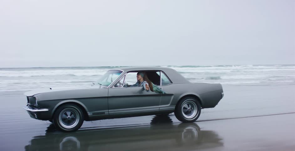 A young woman in a classic grey Mustang enjoys a carefree drive along the ocean shoreline on a moody, overcast day