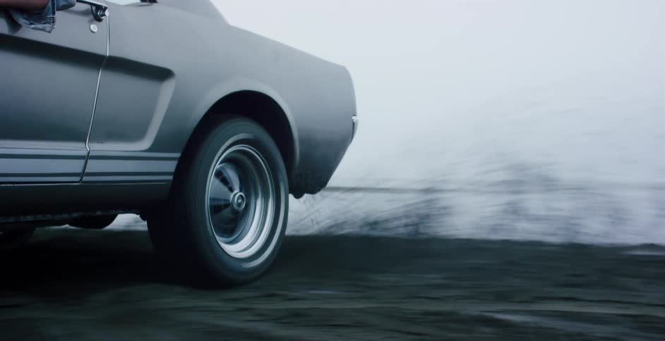 A free-spirited young woman hangs out the window of a classic muscle car as it speeds along a wet, sandy beach on a foggy day