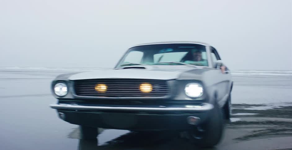 A joyful woman drives her vintage gray Ford Mustang along a wide, foggy beach in slow motion