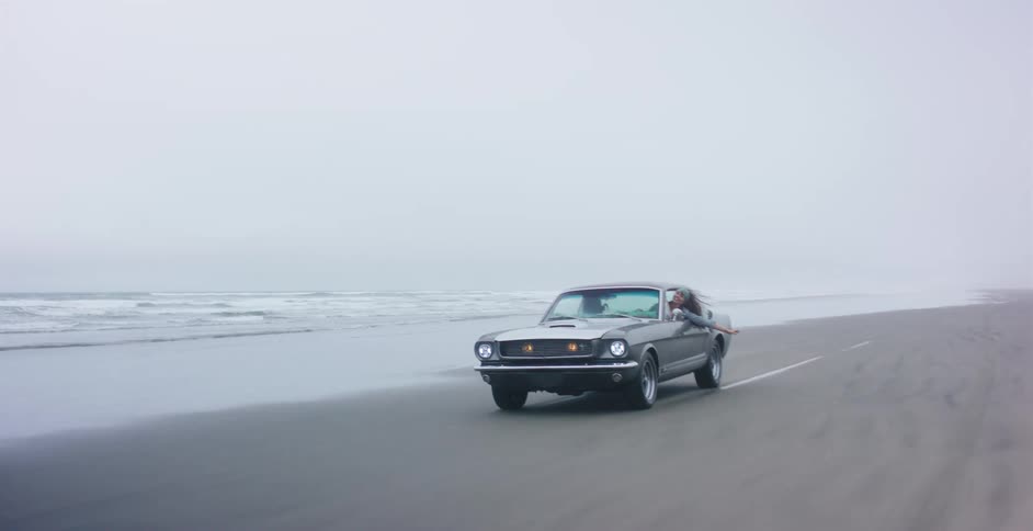 A free-spirited woman hangs out of the window of her classic muscle car while driving along the beach on a misty day