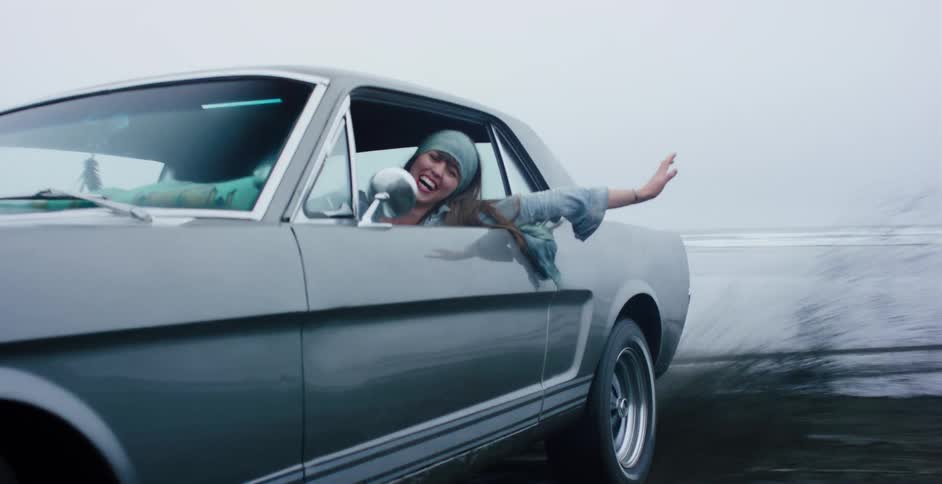 A carefree woman smiles and waves from the driver's window of a vintage muscle car as it speeds along a foggy beach in slow motion