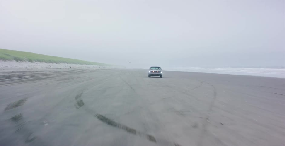 A vintage car with its headlights on drives down a vast, foggy beach next to the ocean shoreline