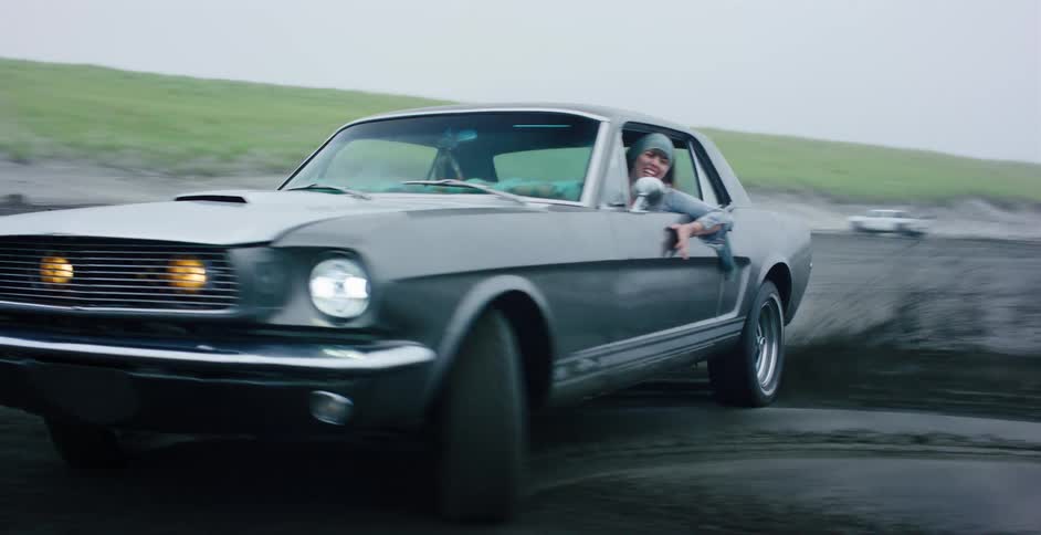 A joyful young woman smiles while driving a classic grey Ford Mustang in slow motion on a wet, sandy beach on a foggy day