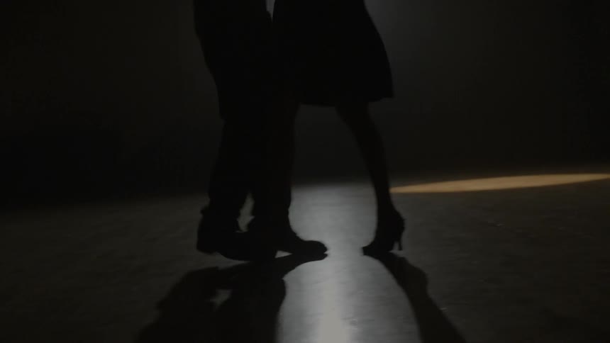 The legs of a man and woman dance on a wood floor lit with only one spotlight