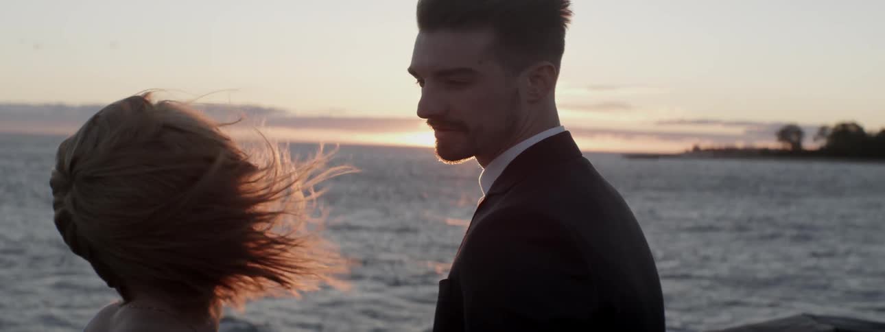 A man and woman stand on a windy beach at sunset