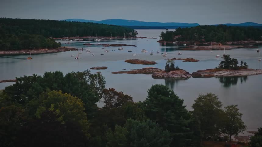 Sailboats rest peacefully in a tranquil, tree-lined harbor dotted with small islands under an overcast sky