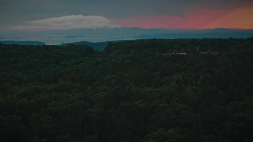 A dramatic sunset paints the sky with pink and orange hues over a vast, dark green forest and distant misty mountains