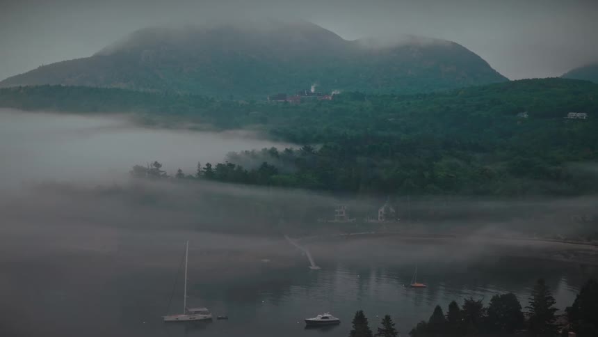 A misty harbor filled with sailboats rests quietly at the base of a large, forested mountain