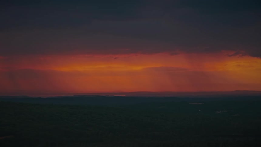 Storm clouds part to reveal a vibrant sunset over a dark, rolling landscape
