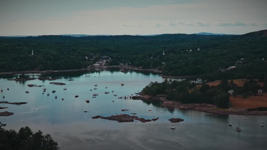 A serene, high-angle view of sailboats anchored in a calm harbor surrounded by dense forests and a quaint coastal town