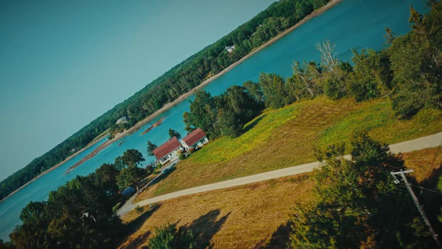 A tilted aerial shot flies over a lush green forest towards a house on the bank of a wide river on a sunny day