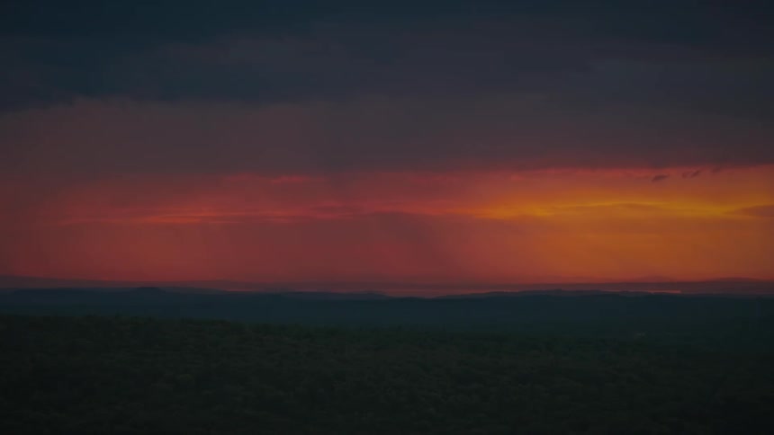 A vivid sunset of red and orange glows through dark storm clouds as rain falls over a silhouetted forest