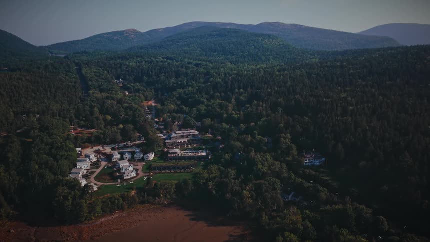 A slow aerial push-in shot reveals a charming coastal town nestled between a vast green forest and a bay at low tide, with rolling hills in the background
