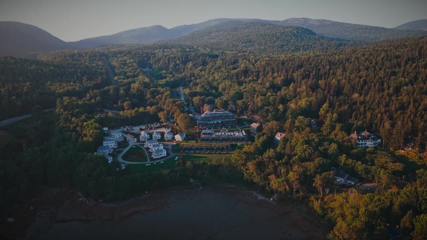 Slowly ascending aerial shot reveals a beautiful coastal resort nestled between a tranquil bay and a vast, mountainous forest during sunrise