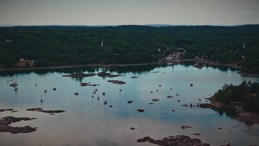 An expansive aerial view of sailboats moored in a calm, reflective bay surrounded by green forests and a small coastal town