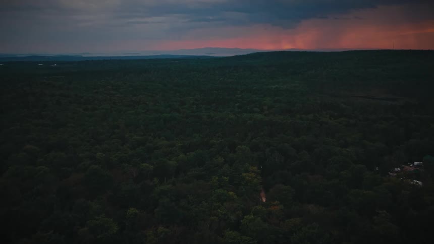 A slow aerial flight over a vast, dark forest at dusk, revealing a dramatic red sky and stormy clouds on the horizon