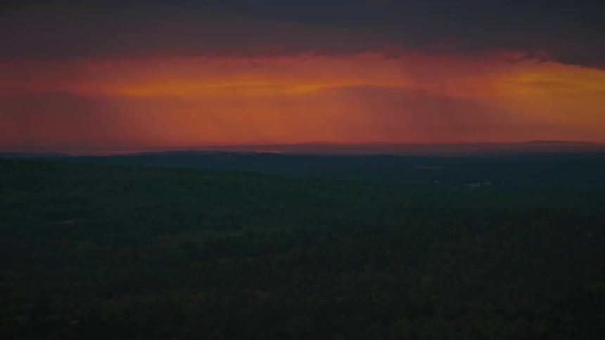 A fiery red and orange sunset glows on the horizon through dark, ominous clouds above a vast, dark forest landscape