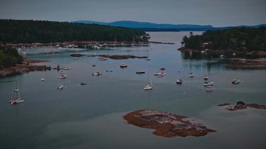 An aerial view slowly advances over a tranquil harbor filled with sailboats and fishing boats anchored along a rocky, forested coastline at dusk