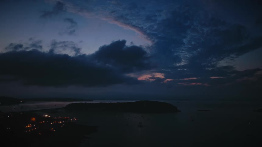 An aerial view of a tranquil coastal town and a silhouetted island under a dramatic and moody twilight sky