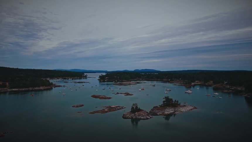 A serene aerial view shows sailboats moored in a calm harbor dotted with rocky islands under a cloudy dusk sky