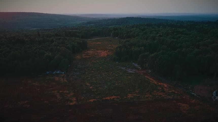 A slow, moody aerial pullback over a harvested field reveals a vast, forested mountain landscape with a cleared pathway at dusk