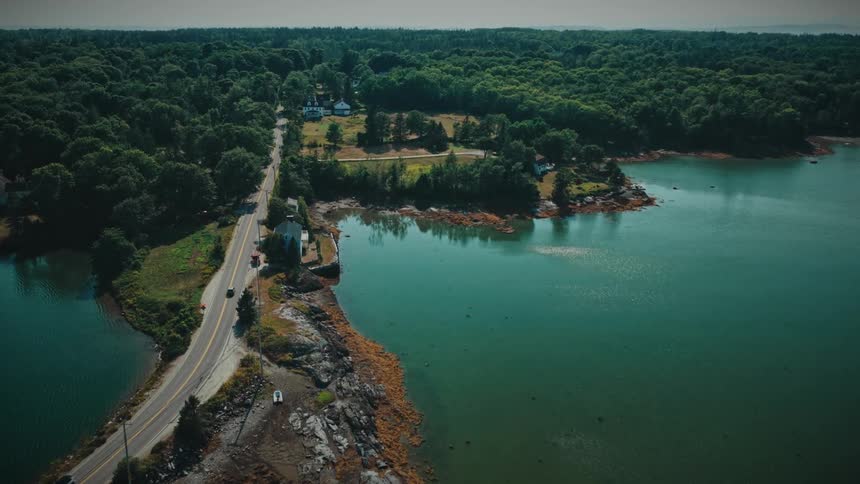 Ascending aerial shot of a scenic coastal road in a lush, forested area next to calm blue-green water on a sunny day
