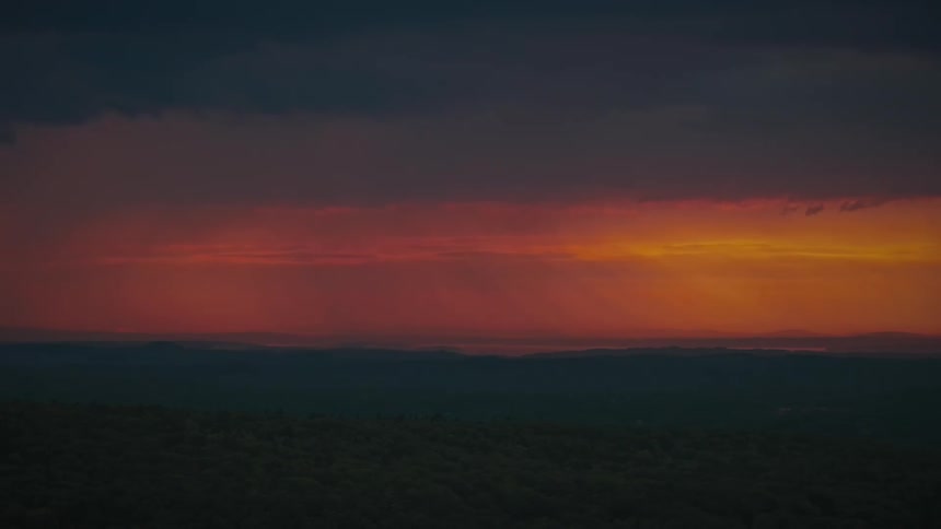 Rain falls from dramatic, fiery red clouds during a beautiful sunset over a silhouetted mountain landscape