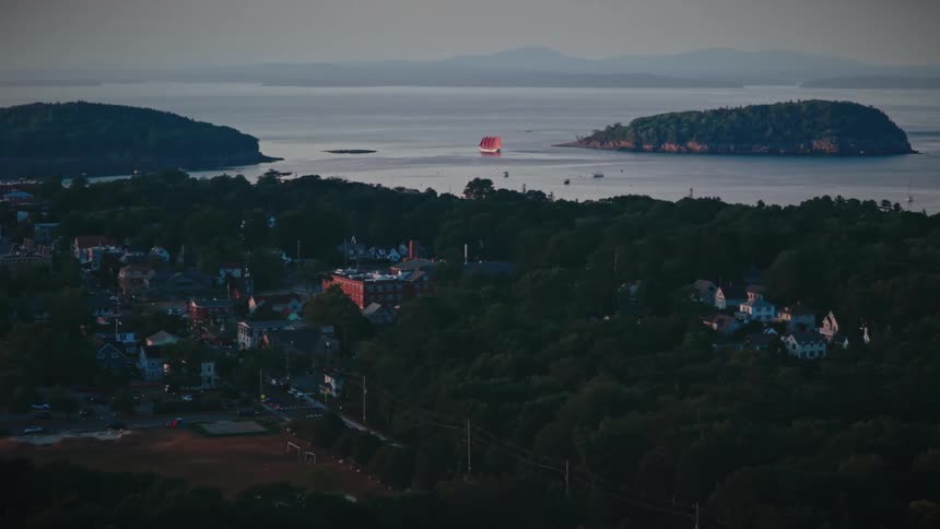 A striking red schooner sails on the ocean past a quiet coastal town and islands at twilight