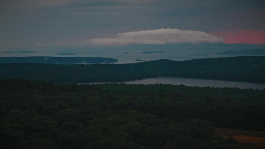 A dramatic aerial shot slowly pulls back over a dense forest to reveal lakes and distant coastal islands under a moody sky at sunset