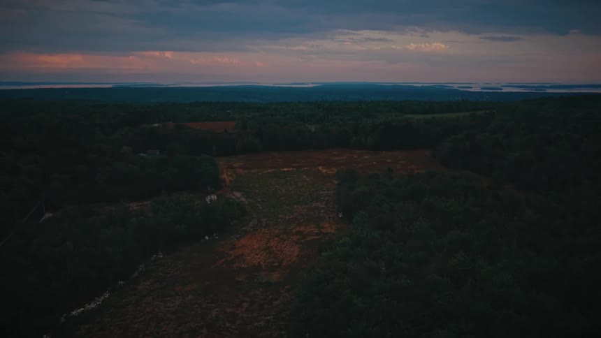 A cinematic aerial view slowly pushes over a vast, dark forest landscape as the last light of a colorful sunset glows on the horizon