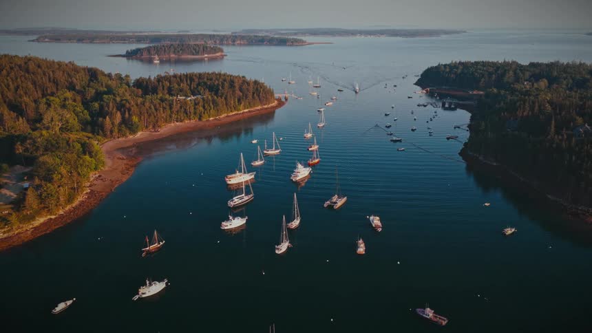 A scenic aerial shot flies over a tranquil harbor packed with sailboats and yachts moored between lush, forested islands at sunset