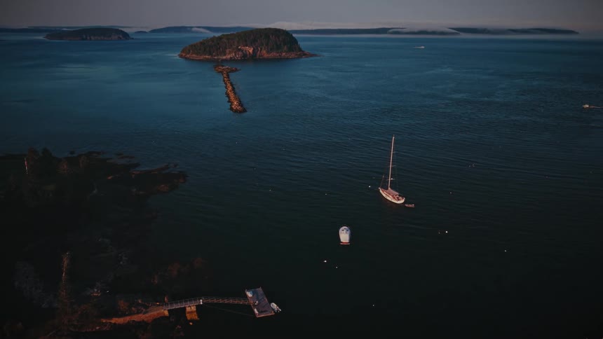 An epic aerial shot pushes over a dense forest towards a calm coastal harbor with sailboats anchored near islands at sunrise