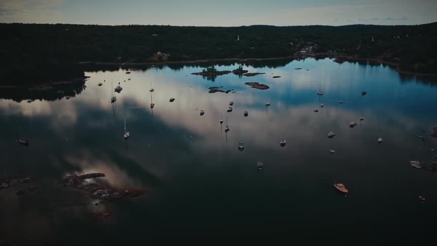A peaceful aerial flight over a tranquil harbor filled with moored sailboats and yachts, with the sky perfectly reflected in the still water