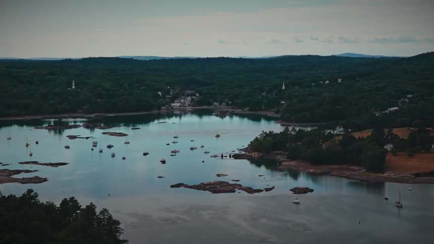 A tranquil aerial view shows sailboats peacefully moored in a secluded harbor, with a charming coastal town nestled in the lush green hills