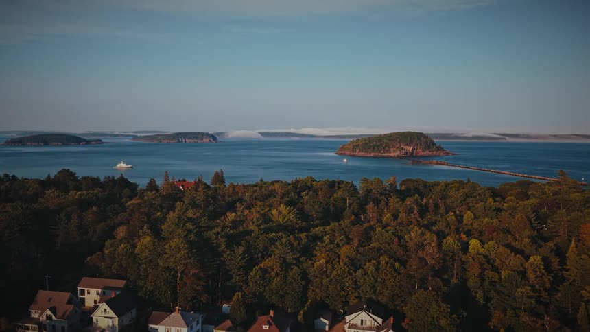 Slow aerial push-in over a tree-covered coastal town towards the beautiful ocean and islands of Bar Harbor, Maine during golden hour