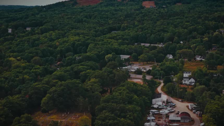 A slow aerial pullback reveals a quaint coastal town with a boatyard and cemetery surrounded by lush green hills and forest