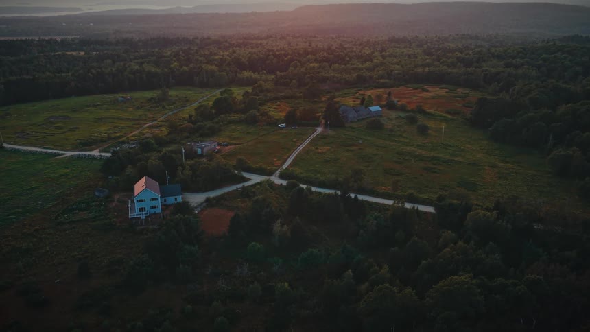 A tranquil aerial shot reveals a secluded country home and barn surrounded by expansive green fields and forests at sunrise