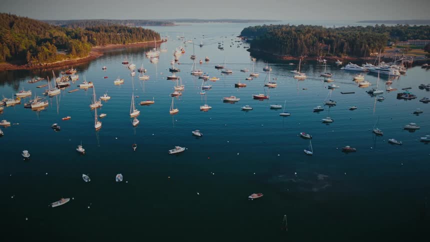 A stunning aerial shot pulls back to reveal a tranquil harbor filled with countless sailboats and yachts nestled along a lush, tree-covered coastline at sunset
