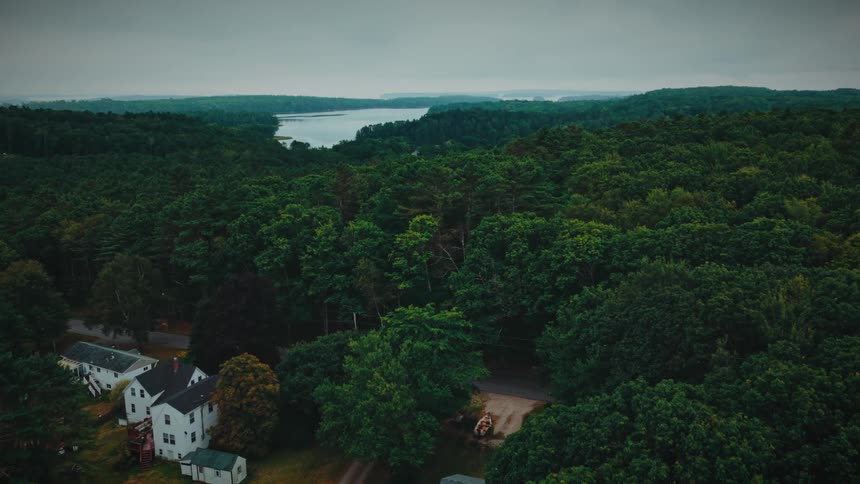 Slow aerial footage moves over a vast green forest with a few rural homes towards a large body of water on a cloudy day