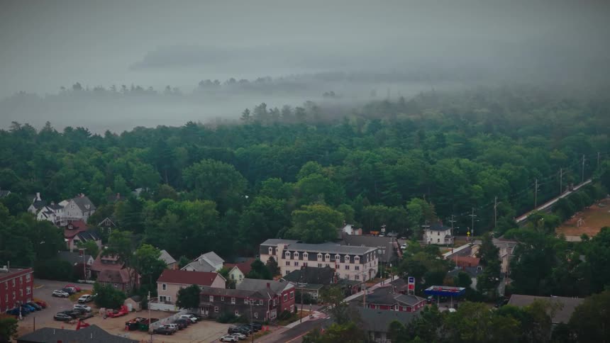 An atmospheric slow zoom over a quaint town surrounded by a vast, foggy forest as a commuter train passes by