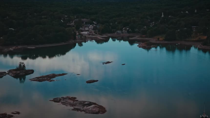 A slow and serene aerial pullback reveals a quiet coastal harbor filled with moored boats surrounded by a dense forest at dusk