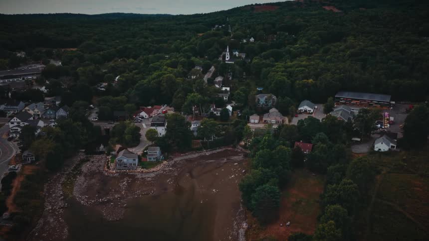 Drone flies over a calm bay towards a small, historic coastal village surrounded by forested hills