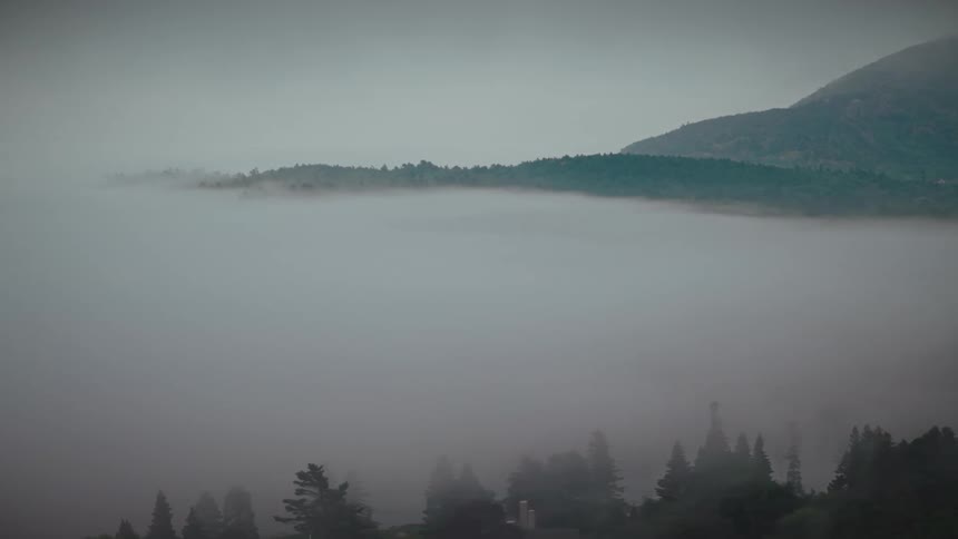 A scenic time-lapse captures a thick blanket of morning fog as it slowly dissipates, revealing a lush green mountain valley and a small town below