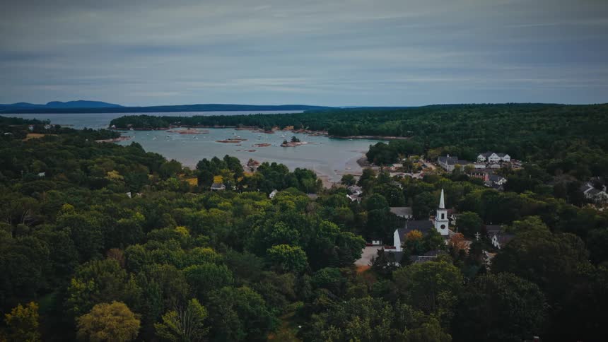 A slow aerial push-in shot over a lush green forest reveals a quaint coastal New England town with a prominent white church steeple and a calm harbor on an overcast day