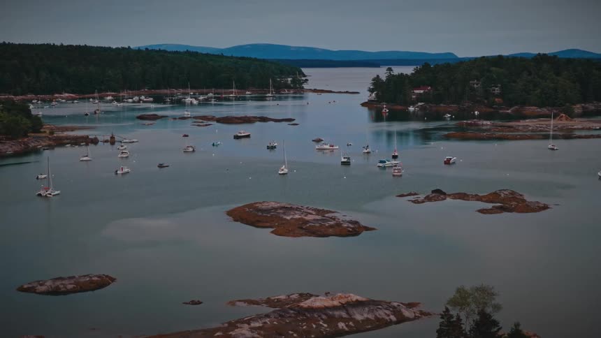 A slow aerial pullback reveals a serene harbor dotted with sailboats and rocky islets along a forested coastline at dusk